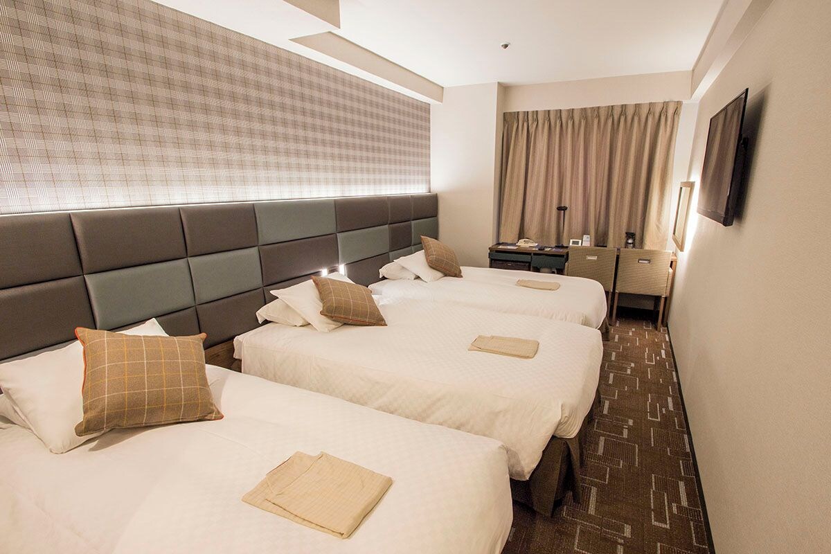 A hotel room with three beds, a large window with curtains, a flat-screen TV, and a desk with a chair.