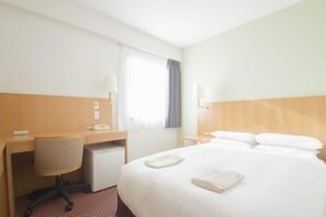 Quarto de hotel com cama, secretária, cadeira, telefone e janela com cortinas.