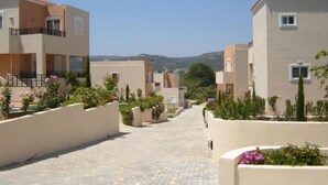 A residential street with modern buildings, a paved walkway, and landscaped areas with plants and flowers. - Selini Villas (Platanias)