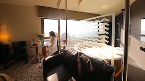 A modern hotel room with a large window, a glass table with fruits and a vase, a leather sofa, and a bathroom with a shower. - Sunrise Hotel (Jounieh)