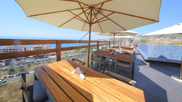 An outdoor terrace with wooden tables and large umbrellas, overlooking a cityscape and the sea. - Sunrise Hotel (Jounieh)