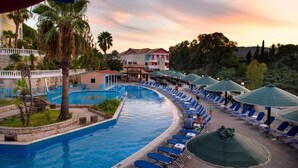 A pool area with lounge chairs and umbrellas, surrounded by palm trees and a building with a red roof. - Zante Imperial Beach Hotel (Zakynthos)