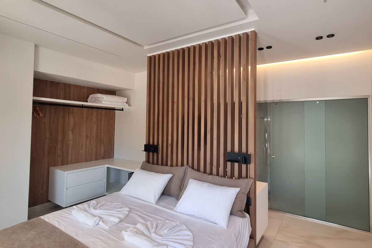 Un dormitorio moderno con una cama grande, un cabecero de listones de madera, un vestidor y un box de ducha de vidrio.