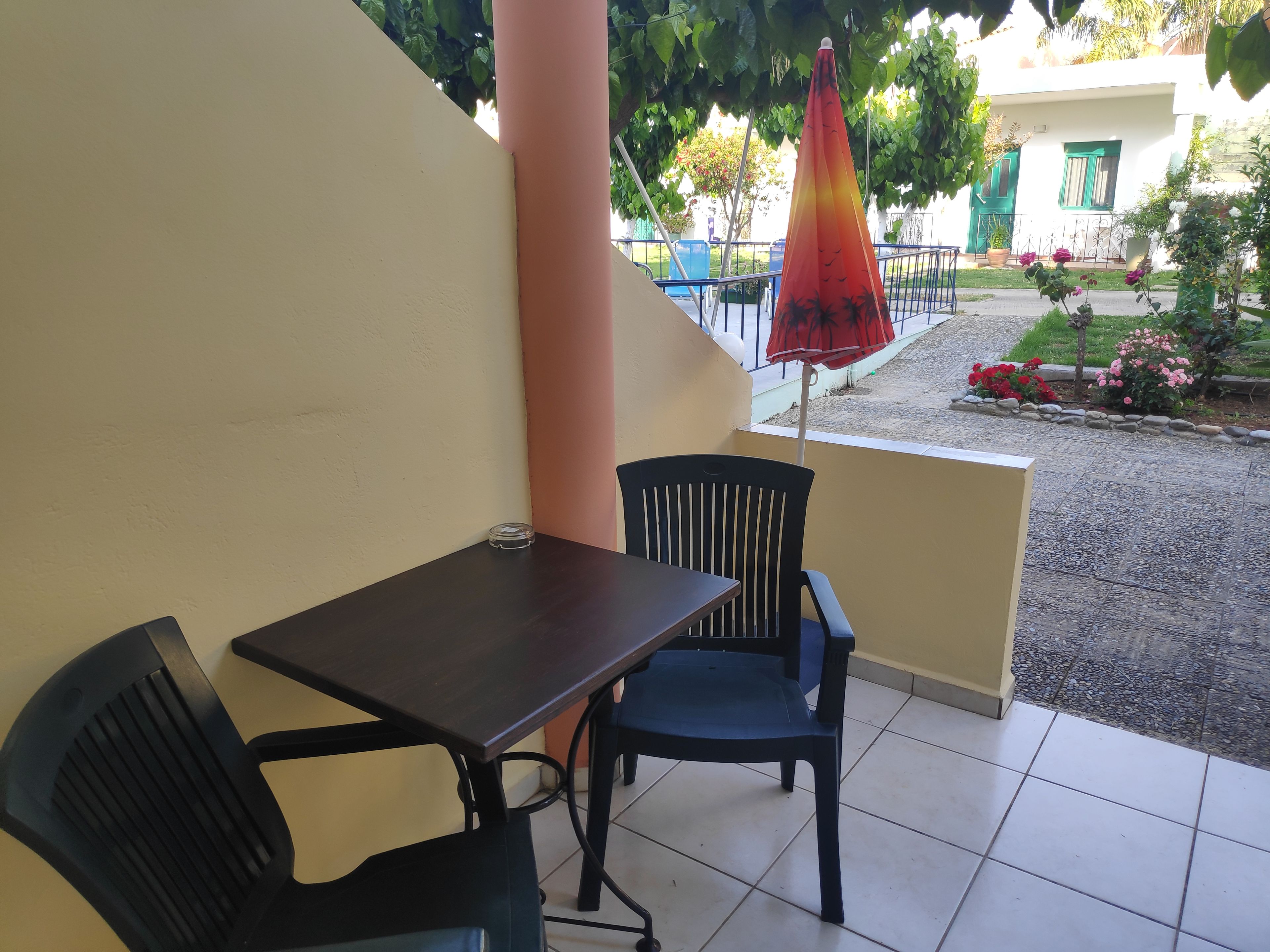 Un patio con una mesa y dos sillas, una sombrilla roja y vistas a un jardín con flores y un edificio al fondo.