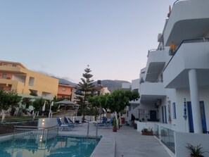 A pool area with lounge chairs, a clear sky, and buildings in the background. - Leonidas Studios & Apartments (Apokoronas)