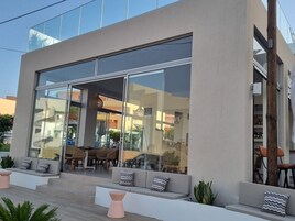 Un edificio moderno con amplios ventanales, asientos exteriores con cojines y un interior visible con muebles y plantas.