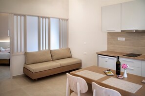 A compact living space with a sofa, a dining table, a kitchen area, and a window with blinds. - Papadakis Apartments (Apokoronas)