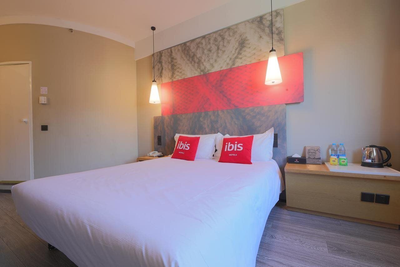 A hotel room with a large bed, two red pillows, a nightstand with a phone, a kettle, and a small table with water bottles.