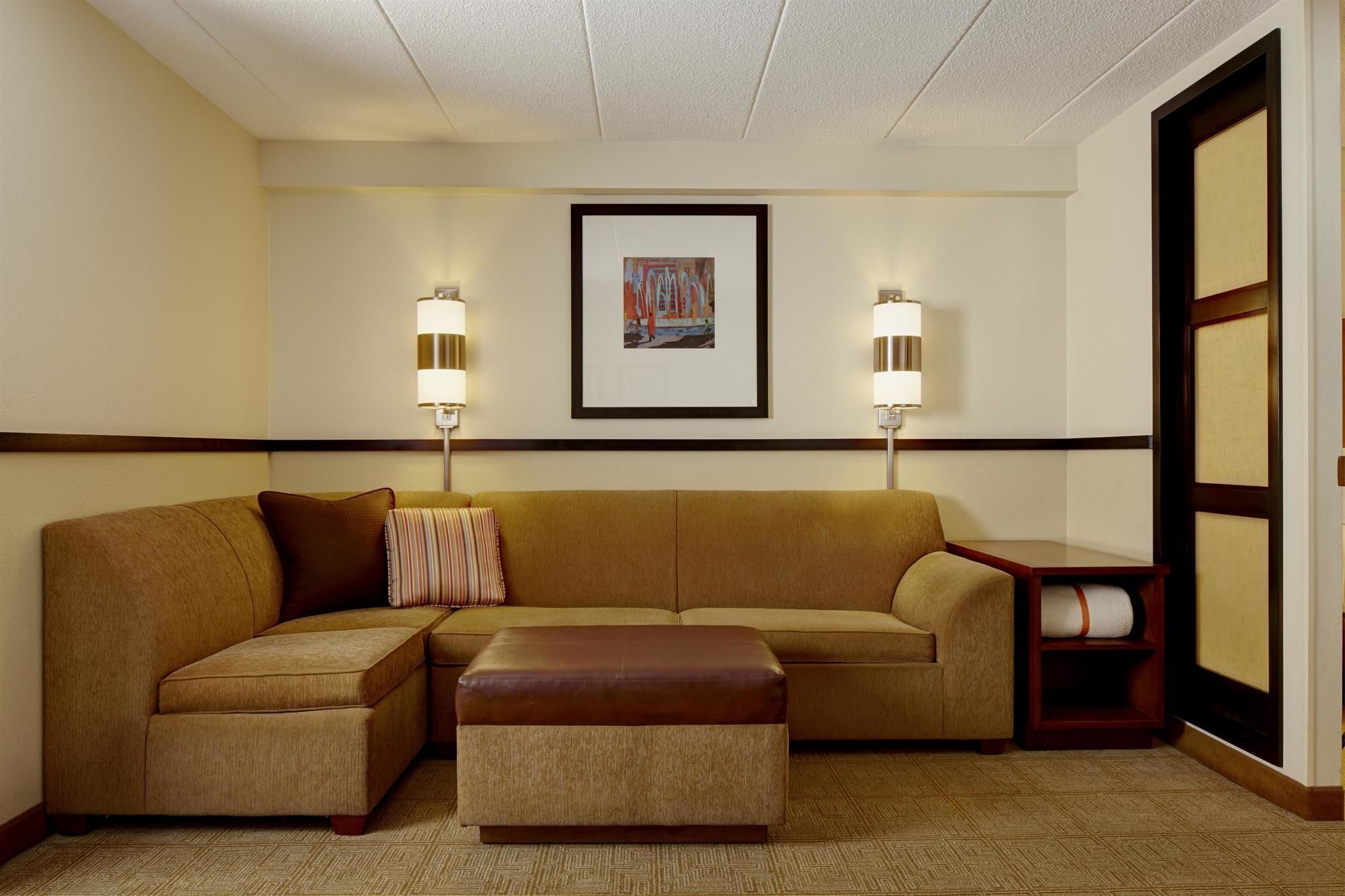 Habitación de hotel con un amplio sofá en forma de L, un reposapiés a juego, dos lámparas de pared y una obra de arte enmarcada sobre el sofá.