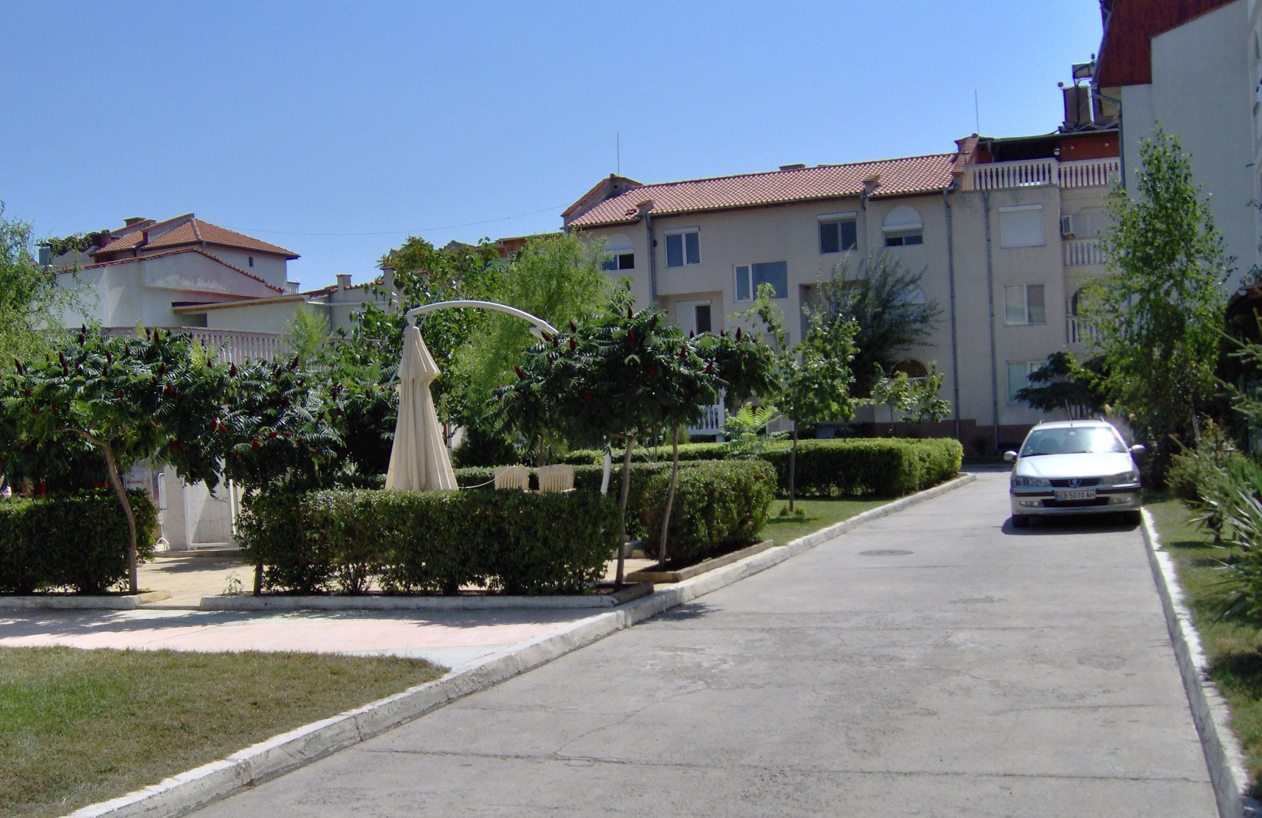 A residential street with a white car parked on the side, a statue in the center, and buildings with red-tiled roofs.