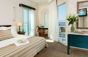 Habitación de hotel con cama, escritorio, silla, cómoda, un jarrón con flores y televisor en la pared.