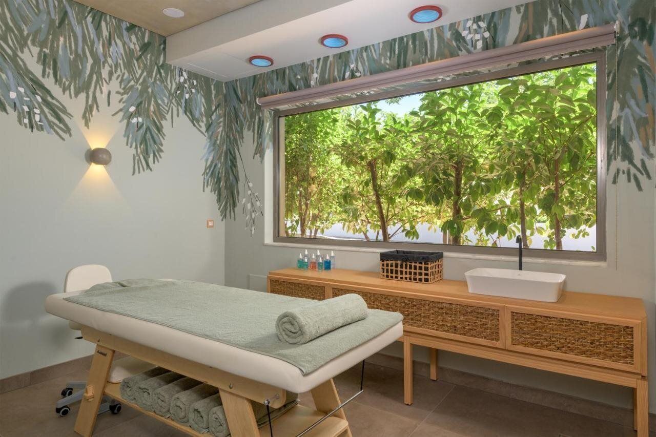 A spa room with a massage table, a wooden bench with a sink, and a large window with a view of trees.