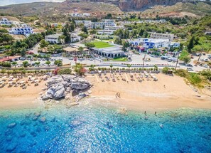 An aerial view of a coastal town with a sandy beach, clear blue waters, and buildings with a Mediterranean architectural style.