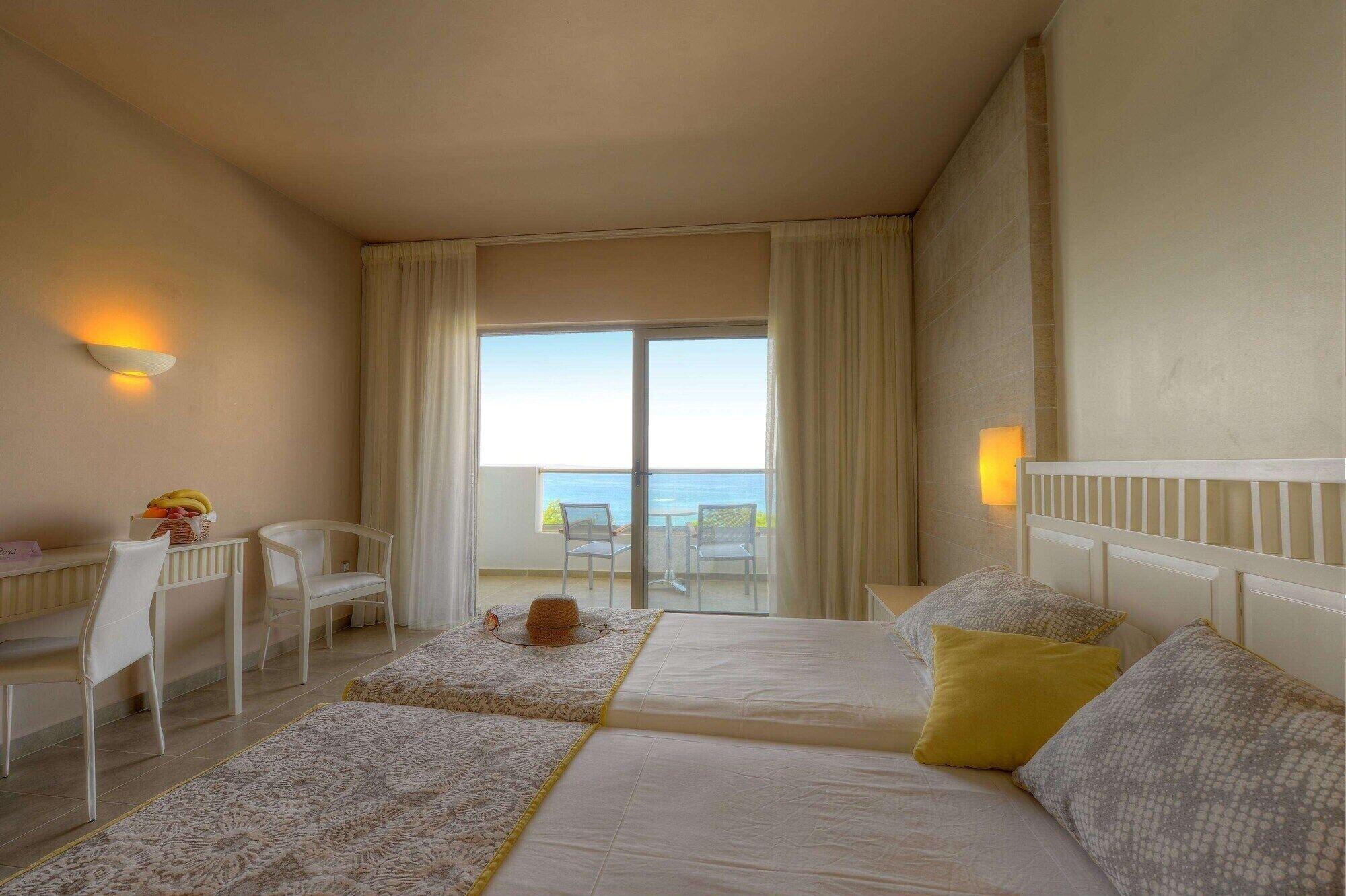 A hotel room with two beds, a desk with a chair, a small table with fruit, and a view of the sea through a sliding door.