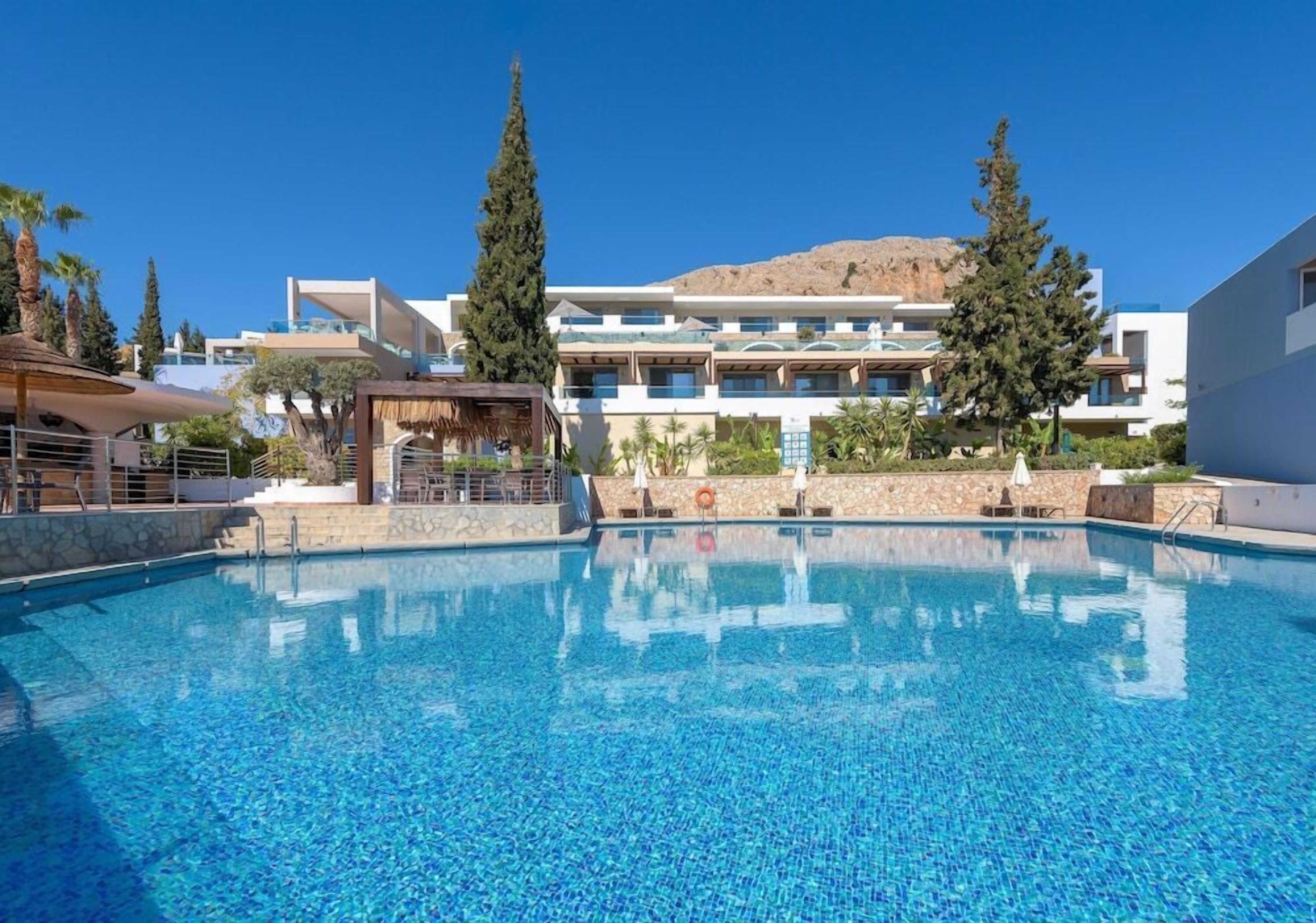 Une piscine à l’eau d’un bleu cristallin, entourée de bâtiments modernes, de palmiers et, au loin, d’une montagne.