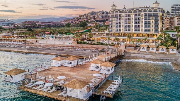A coastal hotel with a pool area, outdoor seating, and a view of the cityscape in the background. - Aria Resort & Spa Hotel (Alanya)