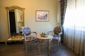 A room with two ornate chairs, a small round table, a framed picture on the wall, and a mirror.