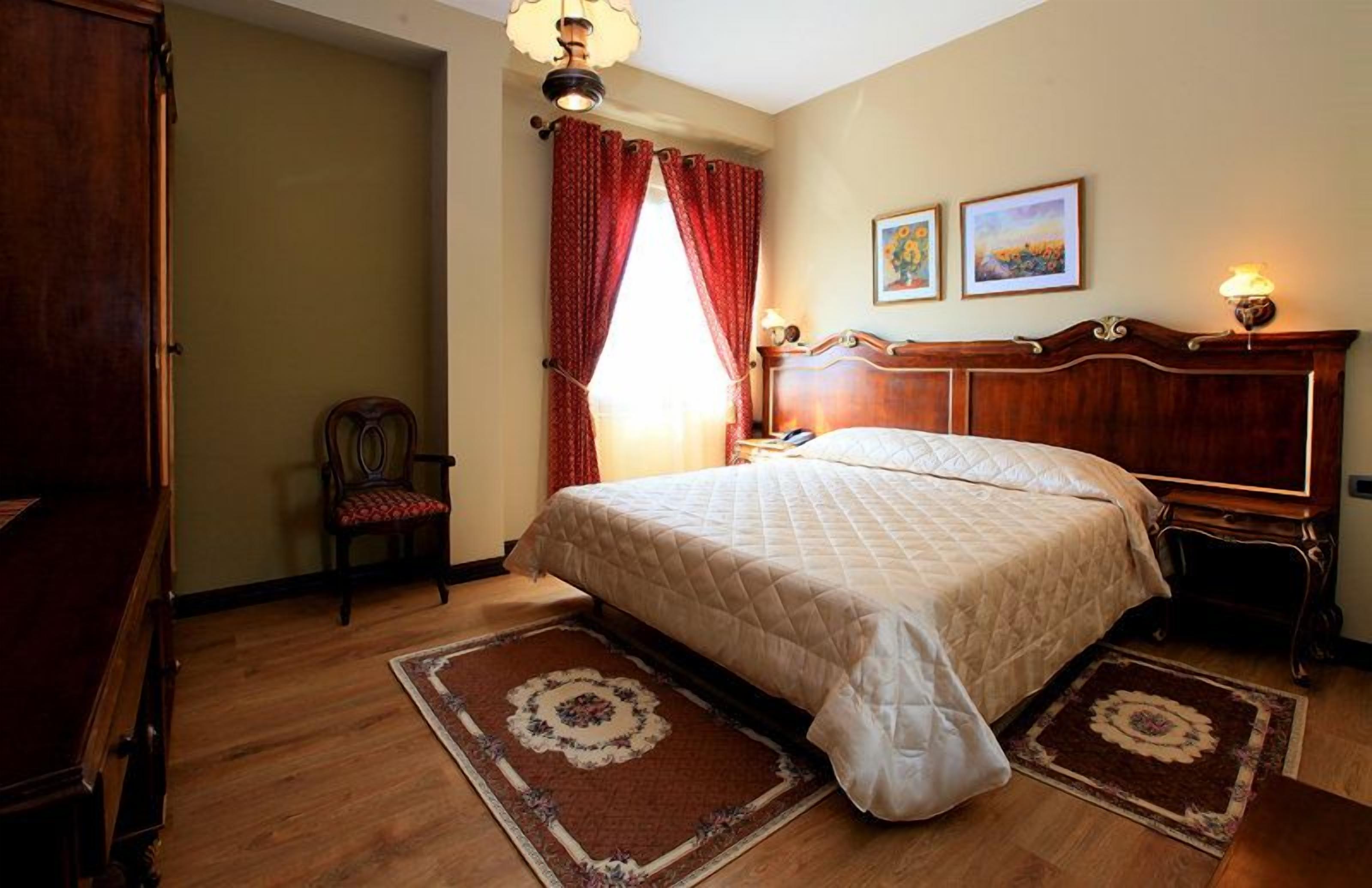 A bedroom with a large bed, a wooden wardrobe, a chair, and a nightstand with a lamp.