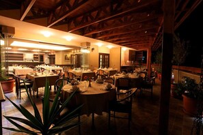 An indoor dining area with tables and chairs, wooden beams on the ceiling, and potted plants.