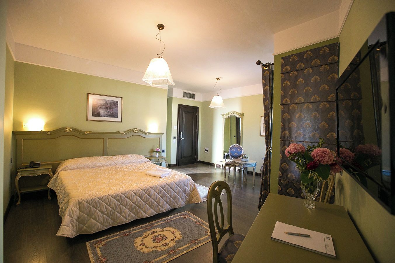 Habitación de hotel con cama, escritorio, silla, espejo y un florero con flores.