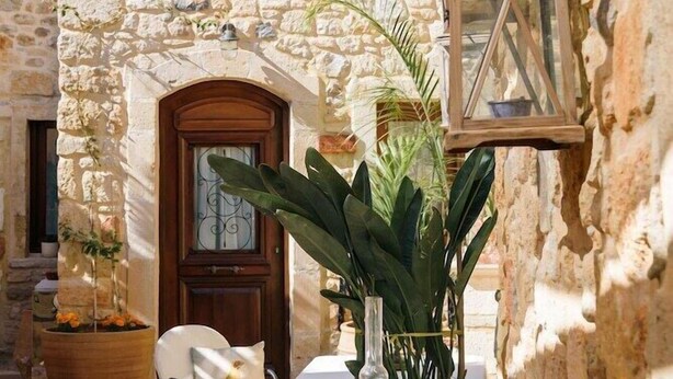 A stone building with a wooden door and windows, outdoor seating with tables and chairs, and potted plants. - Casa Di Veneto (Hersonissos)