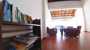 A room with a bookshelf filled with books, a wooden table with chairs, and a view of the outdoors through a window with a wooden ceiling. - Dalawella Beach Resort (Unawatuna)