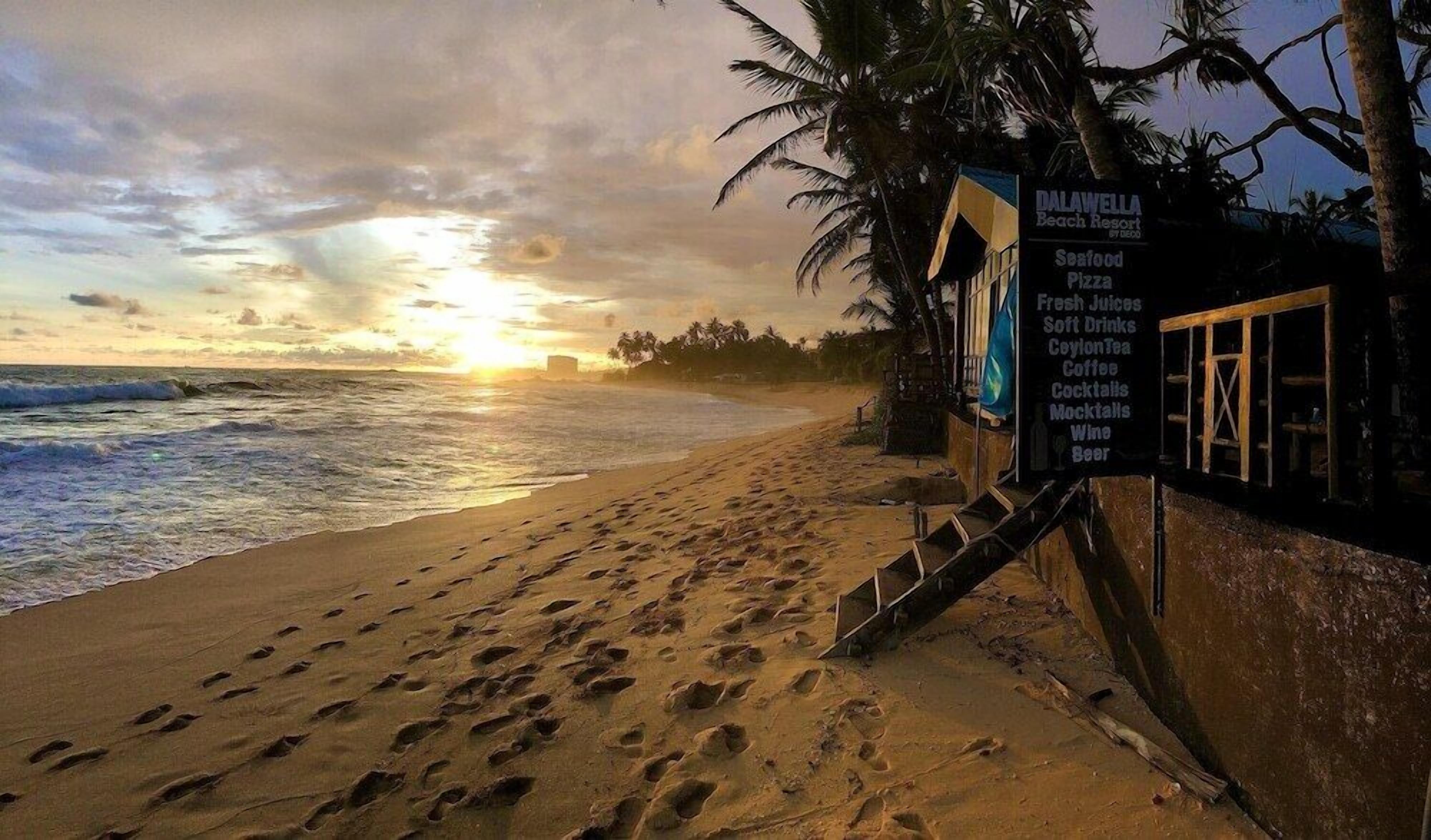 A beach scene with footprints leading to a signboard listing amenities like seafood pizza, fresh juices, and cocktails.
