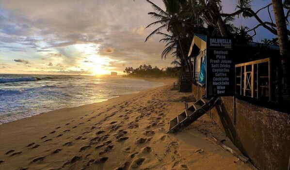 A beach scene with footprints leading to a signboard listing amenities like seafood pizza, fresh juices, and cocktails.