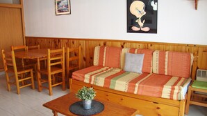 A cozy living room with a wooden sofa, a dining table, and chairs. There is a framed picture on the wall and a cartoon character poster.