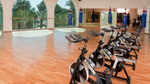 A spacious gym with a row of stationary bikes, large windows with a view of palm trees, and a wooden floor. - Herods Vitalis Spa Hotel Eilat (Eilat)