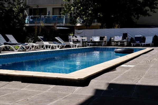 Piscina rectangular con borde de azulejos, rodeada de sillones blancos y una zona cubierta con mesas y sillas.