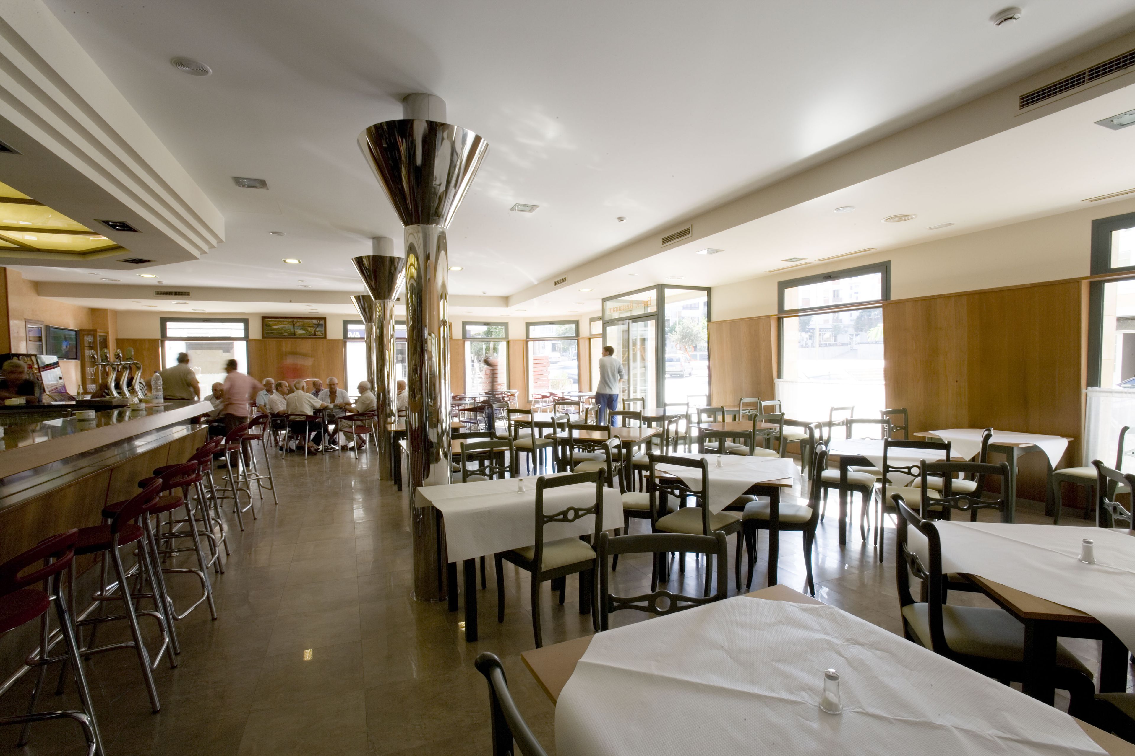 Un restaurante amplio con varias mesas y sillas, una zona de bar y ventanas grandes que permiten la entrada de luz natural.