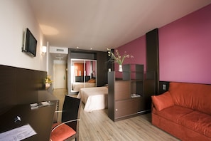 A hotel room with a bed, a desk with a chair, a TV mounted on the wall, and a red sofa.