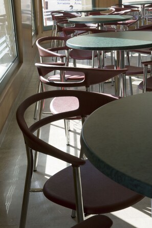 Interior de restaurante con mesas redondas y sillas rojas, ventanas amplias y suelo de baldosas.