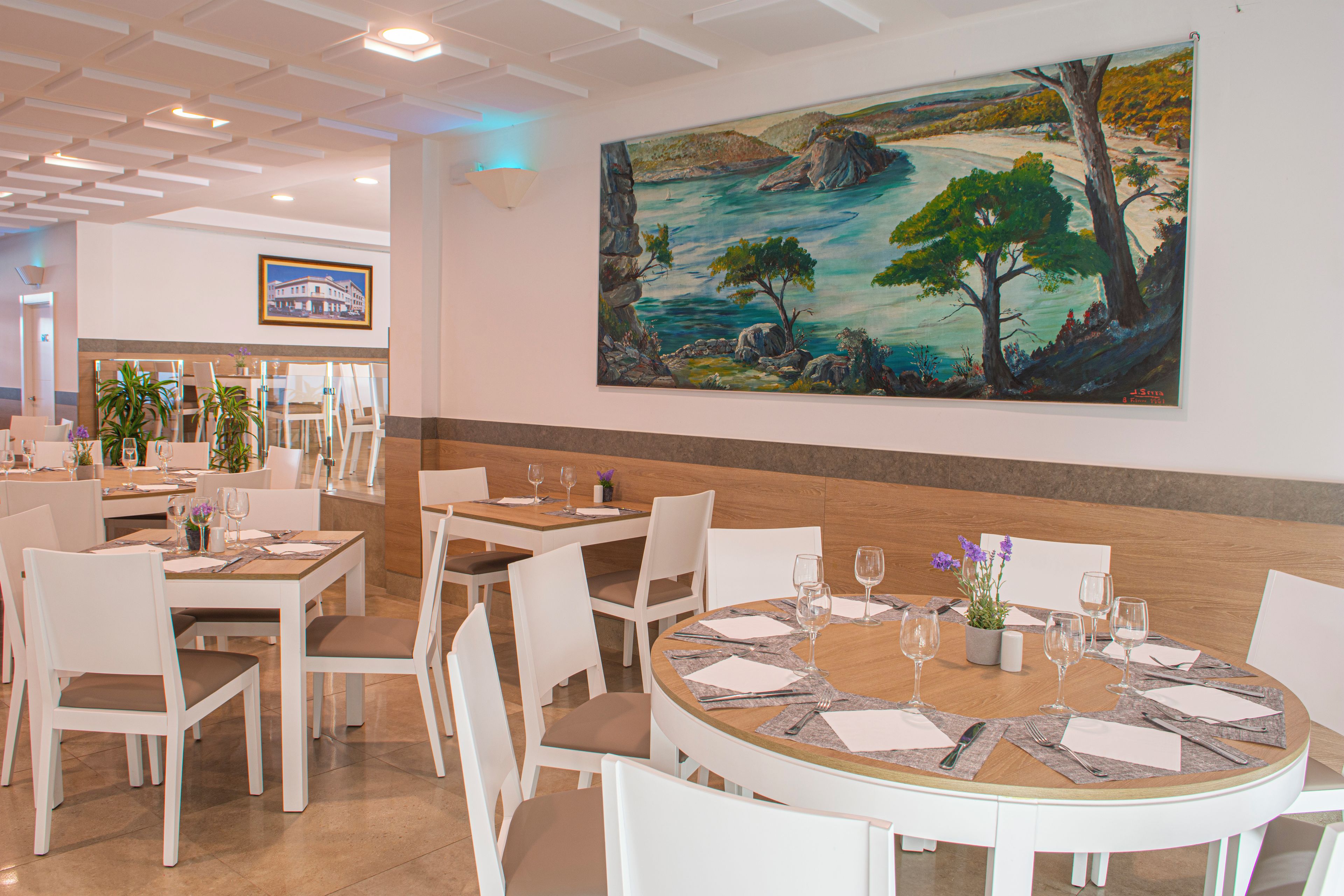 A restaurant with round tables, white chairs, and a large landscape painting on the wall.