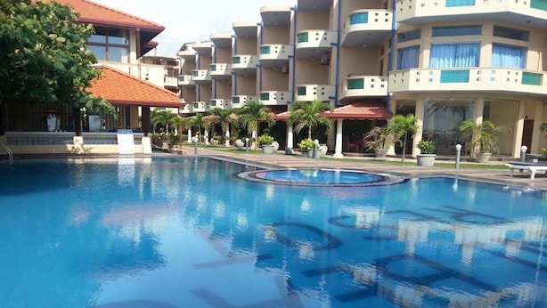 Una piscina rodeada de edificios de varios pisos con balcones, palmeras y una construcción de techo rojo.