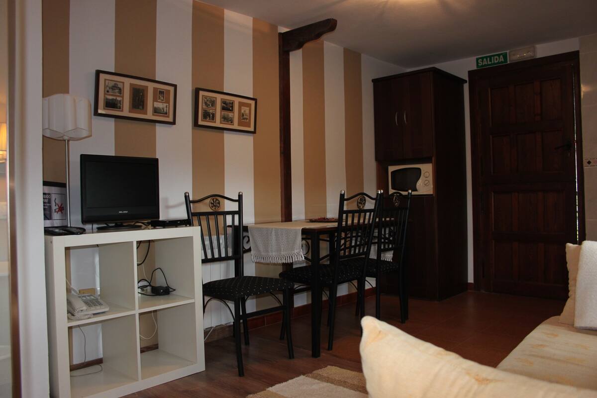 Quarto com paredes listradas, uma televisão sobre um suporte branco, uma escrivaninha com cadeiras, uma cama e uma porta com a inscrição 'SALIDA'.