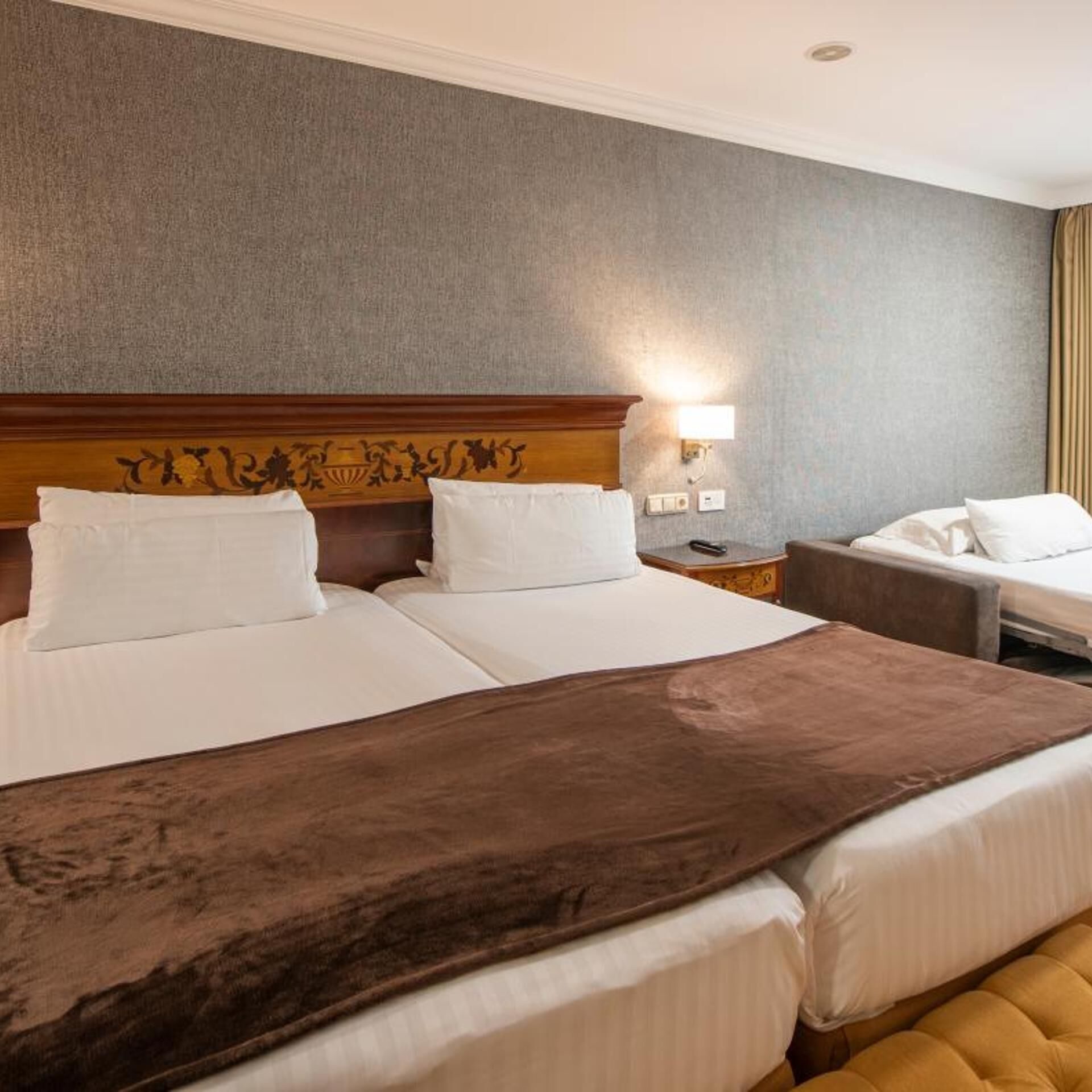 Une chambre d’hôtel avec deux lits, une tête de lit en bois, une table de chevet, une lampe et un banc recouvert d’un tissu marron.