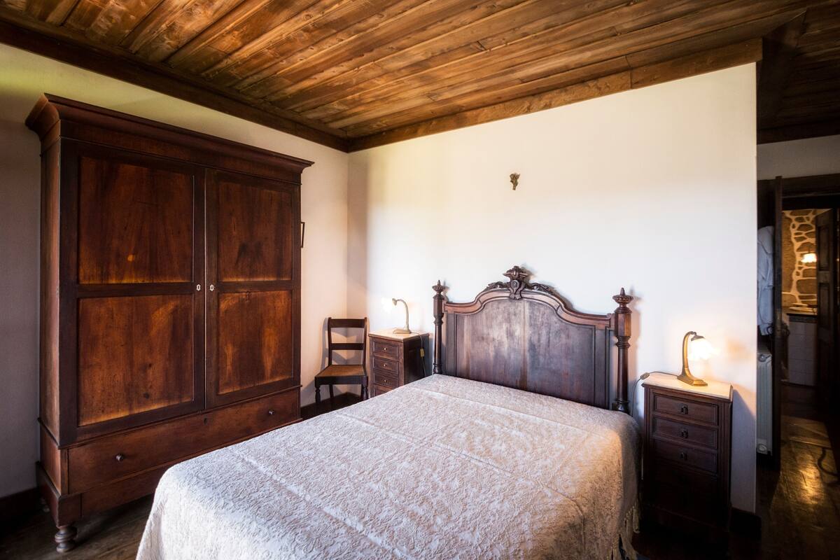 A bedroom with a large wooden wardrobe, a wooden bed with a headboard, two bedside tables with lamps, and a chair.
