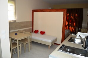 A compact room with a kitchenette, a bed with two red heart-shaped pillows, a small dining table with chairs, and a window with curtains. - Triantafillas Apartments (Rhodes)