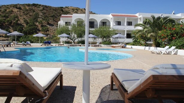 A pool area with lounge chairs and umbrellas, a white building with arches in the background, and a hilly landscape. - Mediterranean Hotel (Karpathos)