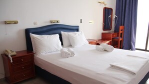 A neatly made bed with white linens and pillows, a wooden nightstand with a telephone, a small wooden dresser, and a mirror on the wall. - Mediterranean Hotel (Karpathos)