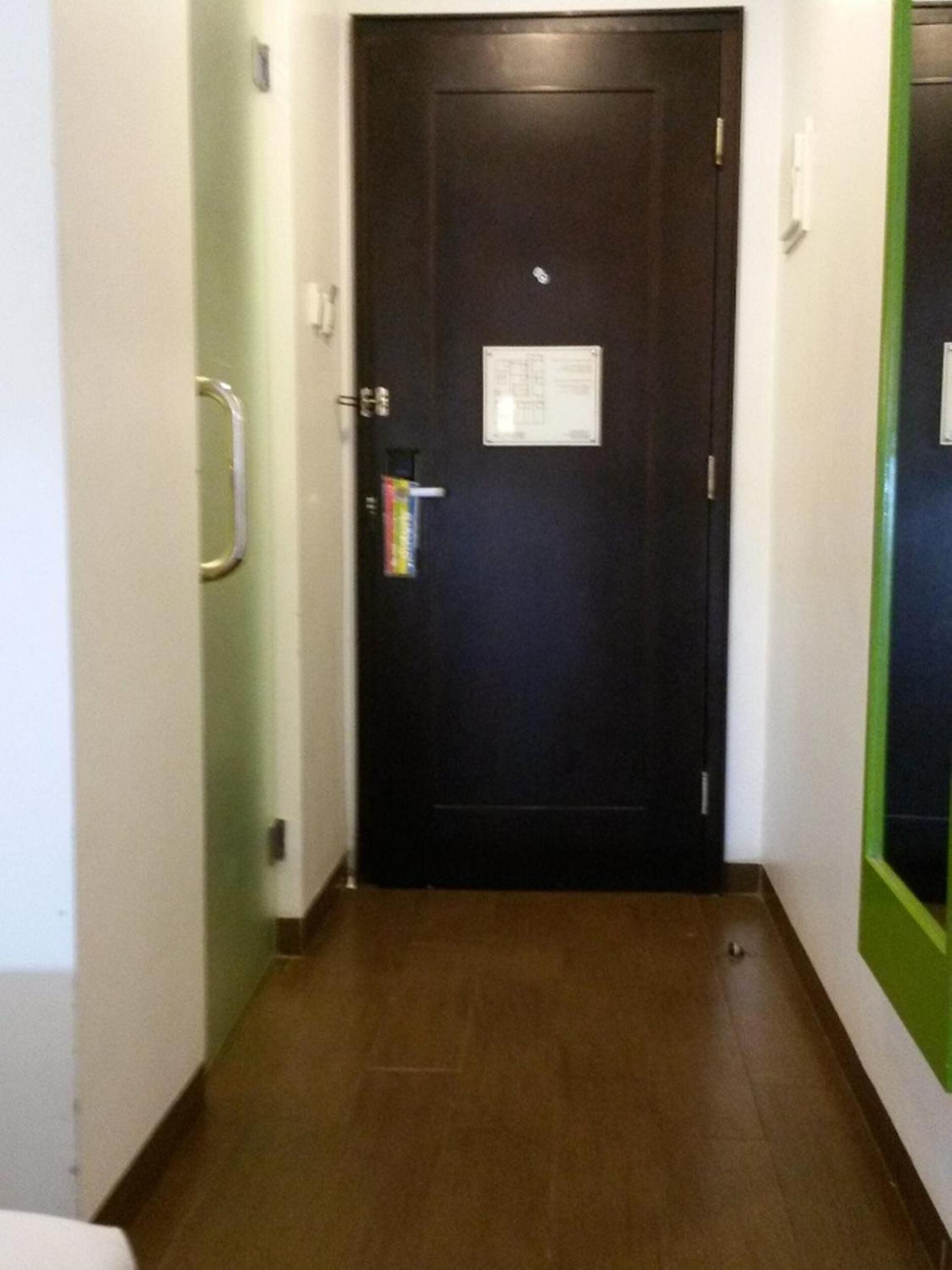 Un couloir étroit avec une porte en bois sombre, une main courante sur le côté gauche et un miroir encadré de vert sur le côté droit.