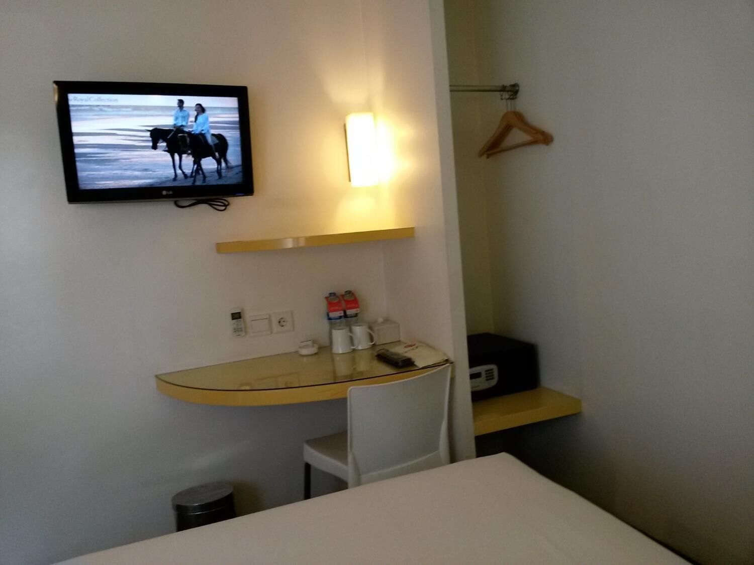 Ett hotellrum med en säng, ett skrivbord med en stol, en tv som är monterad på väggen, ett nattduksbord med en telefon och en kopp, och en klädhängare på väggen.