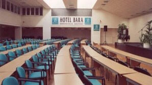 A conference room with rows of blue chairs, a long table, and a podium. A sign above reads "HOTEL BARA." - Bara (Budapest)