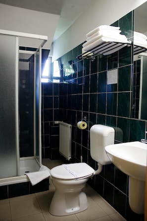 Un baño con inodoro blanco, lavamanos, toallero con toallas dobladas, zona de ducha y pared revestida de baldosas oscuras.
