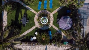 An aerial view of a courtyard with a central circular water feature, surrounded by a stone pathway. There are four blue lounge chairs aligned along the pathway, and a purple umbrella. - Villa Eleni (Corfu)