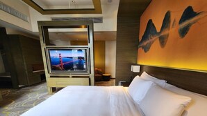 Une chambre d’hôtel avec un grand lit, un téléviseur fixé sur un panneau en bois, un mur orné d’un tableau abstrait et un sol recouvert de moquette.