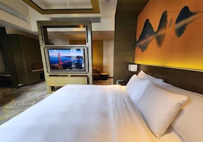 A hotel room with a large bed, a television mounted on a wooden panel, a wall with an abstract painting, and a carpeted floor.