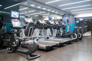 Une rangée de tapis de course et de vélos elliptiques dans une salle de sport, avec des écrans numériques et un mur bleu.
